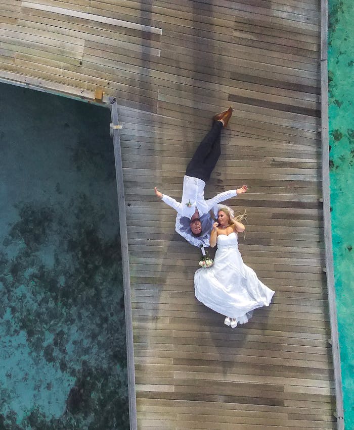 photo-of-bride-and-groom-laying-on-pier-1034356 Aerial view of a bride and groom relaxing on a wooden pier over turquoise waters in the Maldives.
